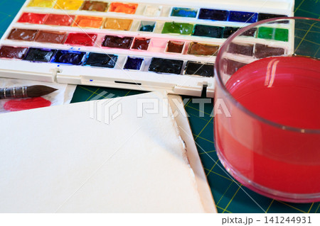 Watercolor paints palette with brush and blank paper sheet, glass jar with red tinted water on green cutting mat background 141244931