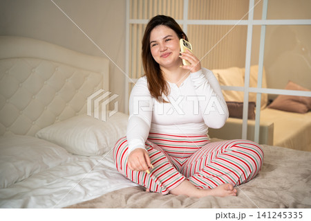 Woman in striped pajamas sitting on bed having a phone conversation at home. Casual home communication and remote consultation lifestyle Woman in striped pajamas sitting on bed having a phone conversation at home. Casual home communication and remote consultation lifestyle 141245335
