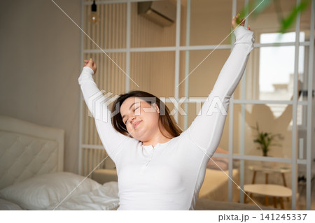 Woman stretching in bed during her morning routine in a cozy bedroom. Sleep wellness relaxation and starting the day with energy Woman stretching in bed during her morning routine in a cozy bedroom. Sleep wellness relaxation and starting the day with energy 141245337