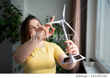 Young woman holding a small wind turbine model indoors near a window. Renewable energy education and green technology concept Young woman holding a small wind turbine model indoors near a window. Renewable energy education and green technology concept 141245342