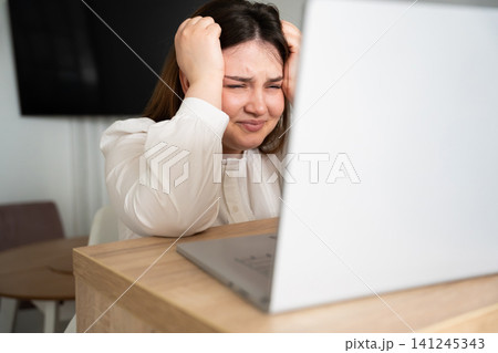 Stressed woman holding her head in front of a laptop at home office. Remote work burnout and deadline pressure in a digital workspace environment Stressed woman holding her head in front of a laptop at home office. Remote work burnout and deadline pressure in a digital workspace environment 141245343