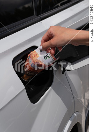 Female hand placing fifty Euro cash note inside vehicle fuel filler neck. Close-up view of white auto gas tank compartment with money symbolizing high petrol prices and rising transportation costs. 141245649