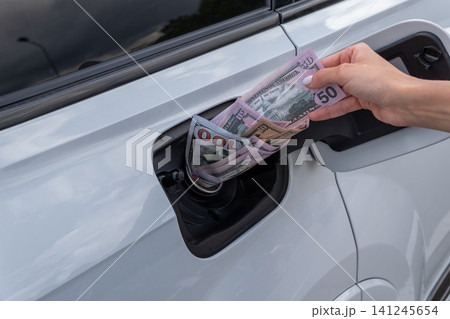 Female hand placing cash money inside vehicle fuel filler neck. Close-up view of white auto gas tank compartment filled with American dollar bills symbolizing high petrol prices and fuel cost. 141245654