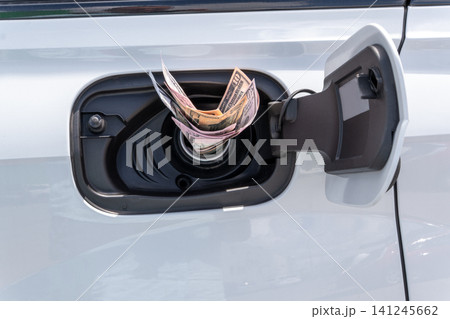 Crumpled cash money inside vehicle fuel filler neck. Close-up view of white auto gas tank compartment filled with American dollar bills symbolizing high petrol prices and expensive transportation. 141245662