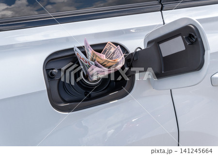 Crumpled cash money inside vehicle fuel filler neck. Close-up view of white auto gas tank compartment filled with American dollar bills symbolizing high petrol prices and expensive transportation. Crumpled cash money inside vehicle fuel filler neck. Close-up view of white auto gas tank compartment filled with American dollar bills symbolizing high petrol prices and expensive transportation. 141245664