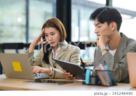 Two Young Asian Business Colleagues Reviewing Documents and Laptop Together at Office Desk 141246440