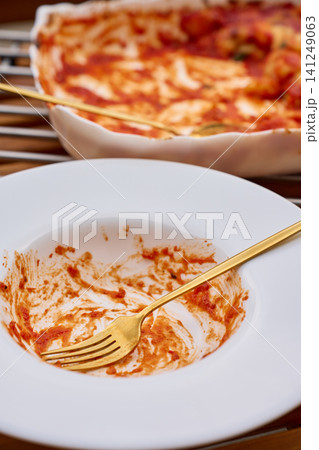 Empty plate with tomato sauce remains and fork in front of baking dish with leftover gnocchi 141249063