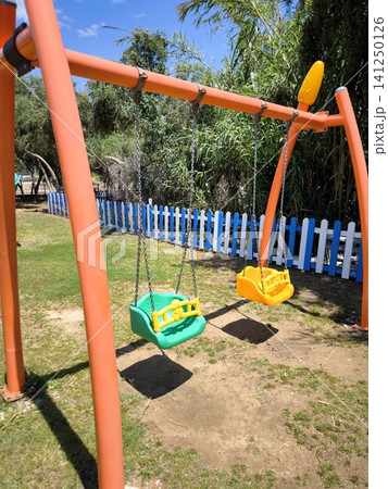 Bright children's swings on a playground in a park, set against a backdrop of greenery. Bright children's swings on a playground in a park, set against a backdrop of greenery. 141250126