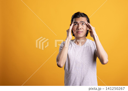 Stressed asian man touching temples with frustrated expression against isolated yellow background. Portrait of young male model massaging forehead, experiencing mental fatigue and tension in studio. 141250267