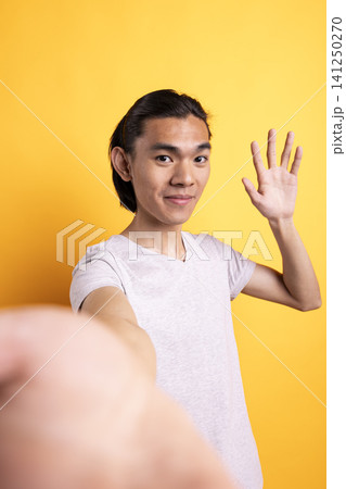 Smiling asian man waving at camera, filming personal vlog in studio. Cheerful male content creator greets online viewers with hello gesture, engaging warmly with social media audience. Smiling asian man waving at camera, filming personal vlog in studio. Cheerful male content creator greets online viewers with hello gesture, engaging warmly with social media audience. 141250270