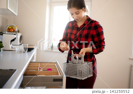 Girl stands in kitchen holding a basket of utensils while sorting items in the drawer in light kitchen 141250552
