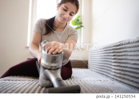 Young girl sits on sofa while cleaning with vacuum cleaner in bright room with sunlight 141250554