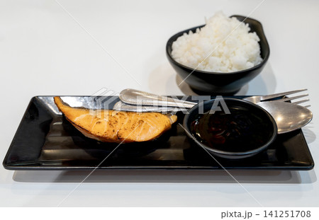 Grilled salmon teriyaki fillet served on a black rectangular plate with a bowl of steamed white rice and soy sauce. Traditional Japanese cuisine meal set isolated on a pure clean white background. Grilled salmon teriyaki fillet served on a black rectangular plate with a bowl of steamed white rice and soy sauce. Traditional Japanese cuisine meal set isolated on a pure clean white background. 141251708