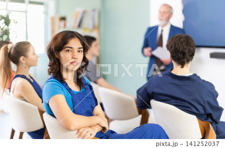 Young woman in blue overalls looking back while sitting in class 141252037