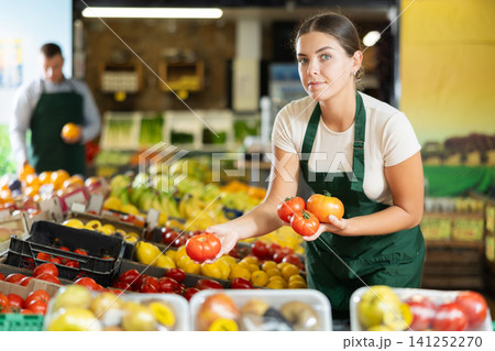 Girl seller of garden stuff store works near showcase, sorting tomato, checking goods 141252270