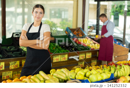 Female seller in uniform standing among shelves with fruits and vegetables and looking in camera Female seller in uniform standing among shelves with fruits and vegetables and looking in camera 141252332