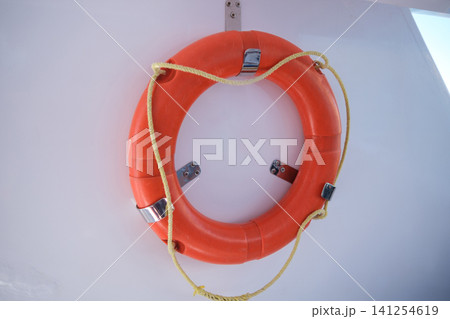 Bright orange life buoy is securely mounted on a white wall, equipped with metal brackets and a yellow rope for marine safety Bright orange life buoy is securely mounted on a white wall, equipped with metal brackets and a yellow rope for marine safety 141254619