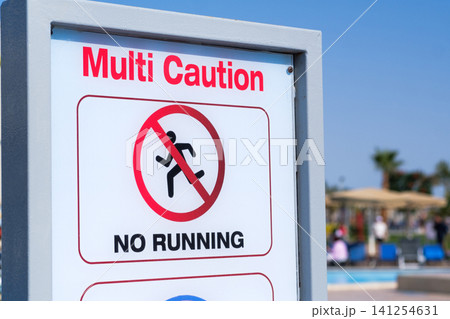 Caution sign indicates "No Running" near a swimming pool, with palm trees and lounge chairs visible in the background under a clear blue sky Caution sign indicates "No Running" near a swimming pool, with palm trees and lounge chairs visible in the background under a clear blue sky 141254631
