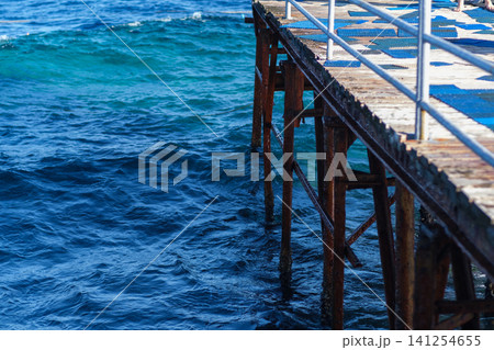 Wooden pier stretches over clear blue ocean water with gentle waves and reflections, illuminated by bright sunlight in a coastal setting 141254655