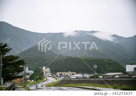 雨が降る曇り空の下で山並みにたなびく雨雲と奥へと続く濡れた道路のある山あいの風景 雨が降る曇り空の下で山並みにたなびく雨雲と奥へと続く濡れた道路のある山あいの風景 141255637