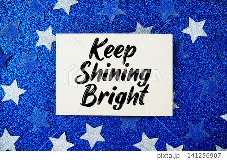 Keep Shining Bright text written on paper card with star glitter on blue background 141256907
