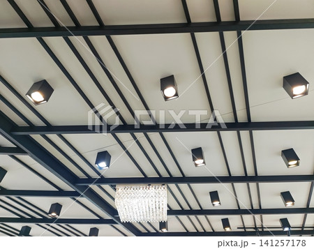 Modern industrial ceiling design with black metal geometric lines, cubic track lights, and a crystal chandelier on a white background. Modern industrial ceiling design with black metal geometric lines, cubic track lights, and a crystal chandelier on a white background. 141257178