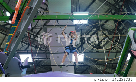 Teenage girl navigating a suspended rope net obstacle high up at an indoor adventure park course. Climbing challenge concept Teenage girl navigating a suspended rope net obstacle high up at an indoor adventure park course. Climbing challenge concept 141258739