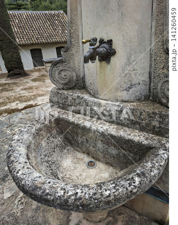 A stone drinking fountain stands in a historic abandoned leprosarium. It features a brass tap and is covered in moss and weathering. The surrounding area shows signs of neglect. A stone drinking fountain stands in a historic abandoned leprosarium. It features a brass tap and is covered in moss and weathering. The surrounding area shows signs of neglect. 141260459