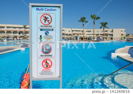 Multi-caution safety sign displayed near a swimming pool, featuring rules for safety, with resort buildings and palm trees visible in the background Multi-caution safety sign displayed near a swimming pool, featuring rules for safety, with resort buildings and palm trees visible in the background 141260658