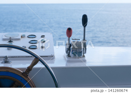 Boat control panel displays throttle lever, steering wheel, and gauges with a view of calm ocean waters in the background Boat control panel displays throttle lever, steering wheel, and gauges with a view of calm ocean waters in the background 141260674
