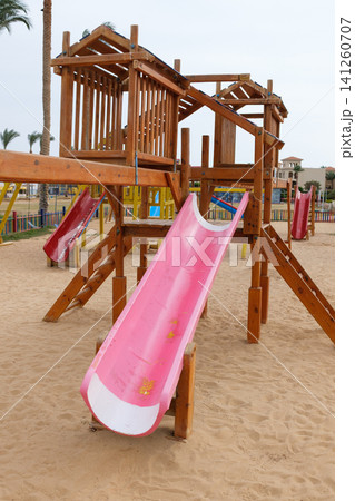 Wooden playground features a pink slide and sandy ground, with colorful play equipment and palm trees visible in the background Wooden playground features a pink slide and sandy ground, with colorful play equipment and palm trees visible in the background 141260707