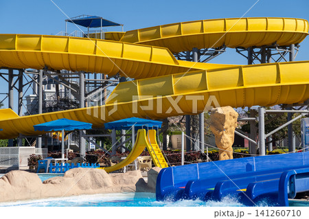 Large yellow water slides twist above a blue pool area in an amusement park, surrounded by sandy areas and umbrellas under a clear sky 141260710
