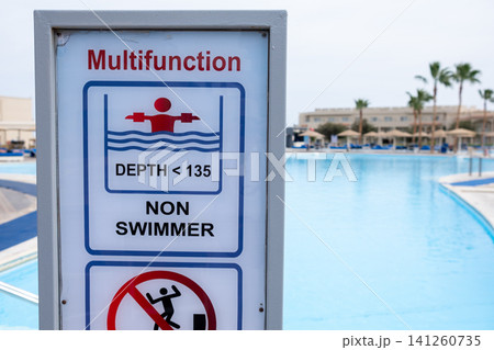 Multifunctional sign at a swimming pool showing depth limit and non-swimmer warning, with a clear blue pool and palm trees visible in the background Multifunctional sign at a swimming pool showing depth limit and non-swimmer warning, with a clear blue pool and palm trees visible in the background 141260735