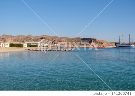 Coastal scene featuring a tranquil beach with clear water, rocky hills in the background, and boats anchored near a small town under a clear sky Coastal scene featuring a tranquil beach with clear water, rocky hills in the background, and boats anchored near a small town under a clear sky 141260741