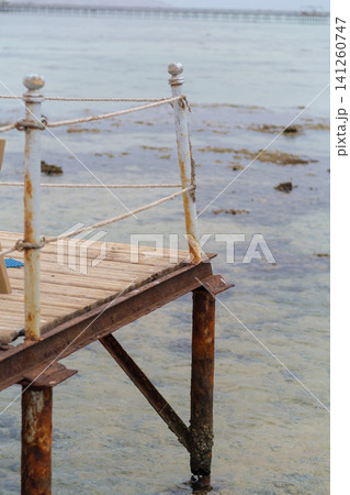Weathered wooden pier with rusted posts and rope railing extends over calm water, revealing a rocky seabed beneath the surface 141260747