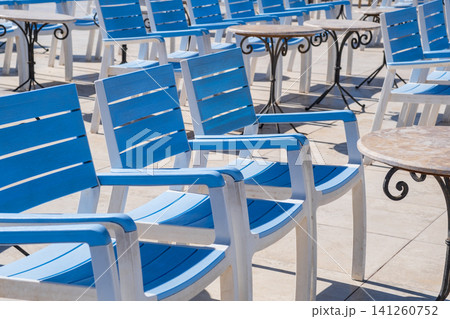 Blue and white outdoor chairs are neatly arranged around round tables on a sunny patio, creating a welcoming space for relaxation and leisure Blue and white outdoor chairs are neatly arranged around round tables on a sunny patio, creating a welcoming space for relaxation and leisure 141260752