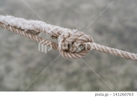 Close-up view of a knot tied in a thick rope, highlighting the intricate details and texture of the fibers against a blurred background 141260763