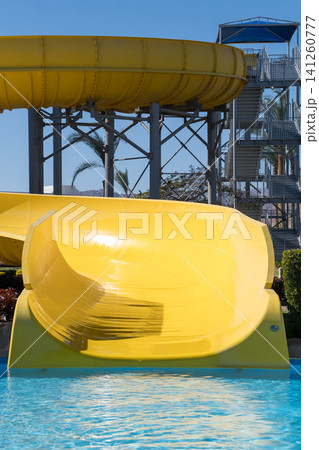Yellow water slide curves into a clear swimming pool, with a tall slide tower and palm trees visible in the background under a bright blue sky Yellow water slide curves into a clear swimming pool, with a tall slide tower and palm trees visible in the background under a bright blue sky 141260777