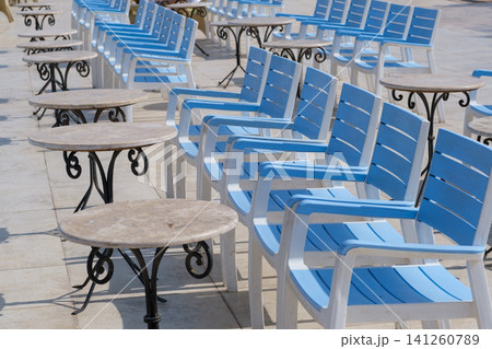 Blue wooden chairs and round tables are neatly arranged on a tiled outdoor patio, illuminated by bright sunlight creating a vibrant scene Blue wooden chairs and round tables are neatly arranged on a tiled outdoor patio, illuminated by bright sunlight creating a vibrant scene 141260789