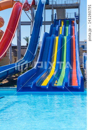 Colorful water slides are positioned next to a clear blue pool at a water park, with a sunny sky and palm trees visible in the background 141260791