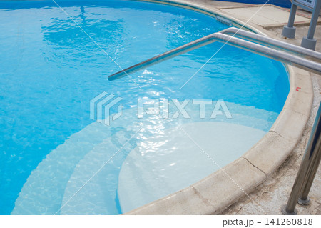 Swimming pool displays clear blue water with a stainless steel handrail, circular step area, and textured concrete edge visible around the perimeter Swimming pool displays clear blue water with a stainless steel handrail, circular step area, and textured concrete edge visible around the perimeter 141260818