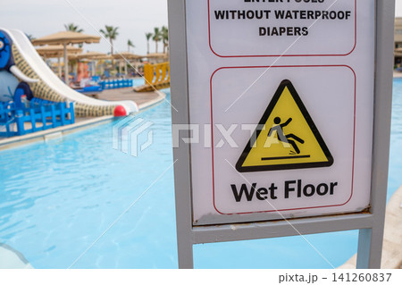 Wet floor sign positioned by a swimming pool featuring a water slide and lounge chairs, set in a resort environment with palm trees 141260837