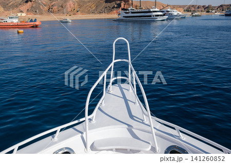 Bow of a white yacht features a railing, with calm blue waters and several boats anchored near a sandy beach in the background Bow of a white yacht features a railing, with calm blue waters and several boats anchored near a sandy beach in the background 141260852