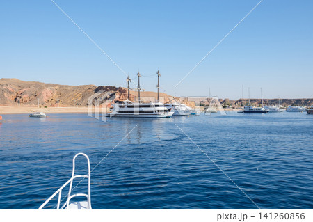 Scenic view featuring multiple yachts and boats anchored in tranquil waters near a rocky coastline with a clear blue sky above Scenic view featuring multiple yachts and boats anchored in tranquil waters near a rocky coastline with a clear blue sky above 141260856