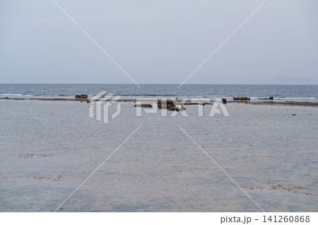 Coastal scene showing remnants of a shipwreck on a shallow beach, calm waters extending to the horizon under a cloudy sky Coastal scene showing remnants of a shipwreck on a shallow beach, calm waters extending to the horizon under a cloudy sky 141260868