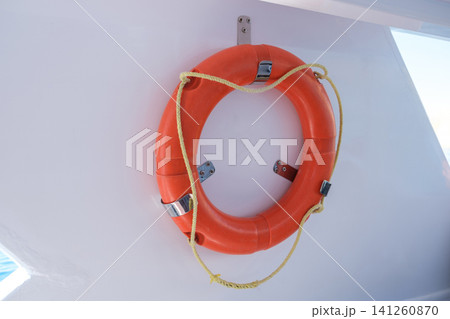 Bright orange life buoy is securely mounted on a white wall, intended for safety on boats and marine vessels in sunny outdoor conditions 141260870