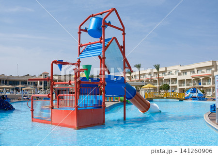 Colorful water play structure features slides and fountains in a resort pool area, with palm trees and hotel buildings in the background Colorful water play structure features slides and fountains in a resort pool area, with palm trees and hotel buildings in the background 141260906