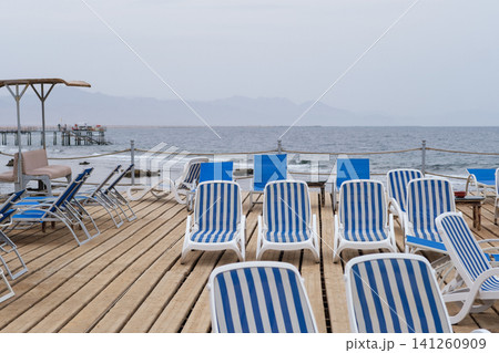 Beachside deck features striped lounge chairs on wooden planks, with calm ocean waters and distant mountains visible in the background Beachside deck features striped lounge chairs on wooden planks, with calm ocean waters and distant mountains visible in the background 141260909