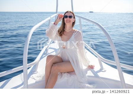 Woman in white beach cover-up relaxes on the bow of a boat, wearing sunglasses, with calm ocean waters and a clear sky in the background Woman in white beach cover-up relaxes on the bow of a boat, wearing sunglasses, with calm ocean waters and a clear sky in the background 141260921