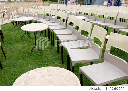 Empty outdoor chairs and tables are arranged neatly on green grass in a public area, prepared for an upcoming event or gathering Empty outdoor chairs and tables are arranged neatly on green grass in a public area, prepared for an upcoming event or gathering 141260932